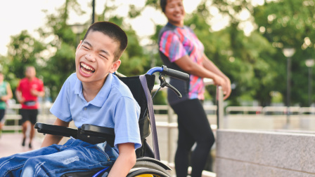 A laughing boy sits in a wheelchair in a school ground. A female adult is in the background, also smiling at the camera. 