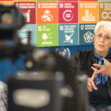 A woman sitting in front of the SDG logos, filmed by a camera