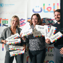 A group of three persons smiling for a photo, holding up cards that indicate their language competencies