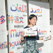 A woman standing in front of a backdrop reading "multilingualism" in different languages. She holds three cards reading "French", "English" and "Chinese" in their respective languages