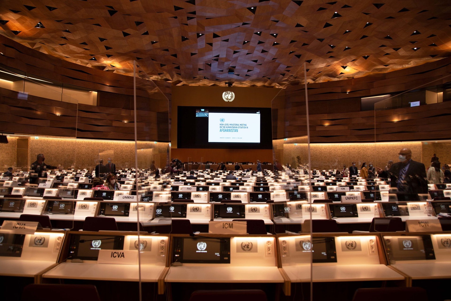 An almost empty conference room, some people are searching seats. A big screen in front shows the title of the event,