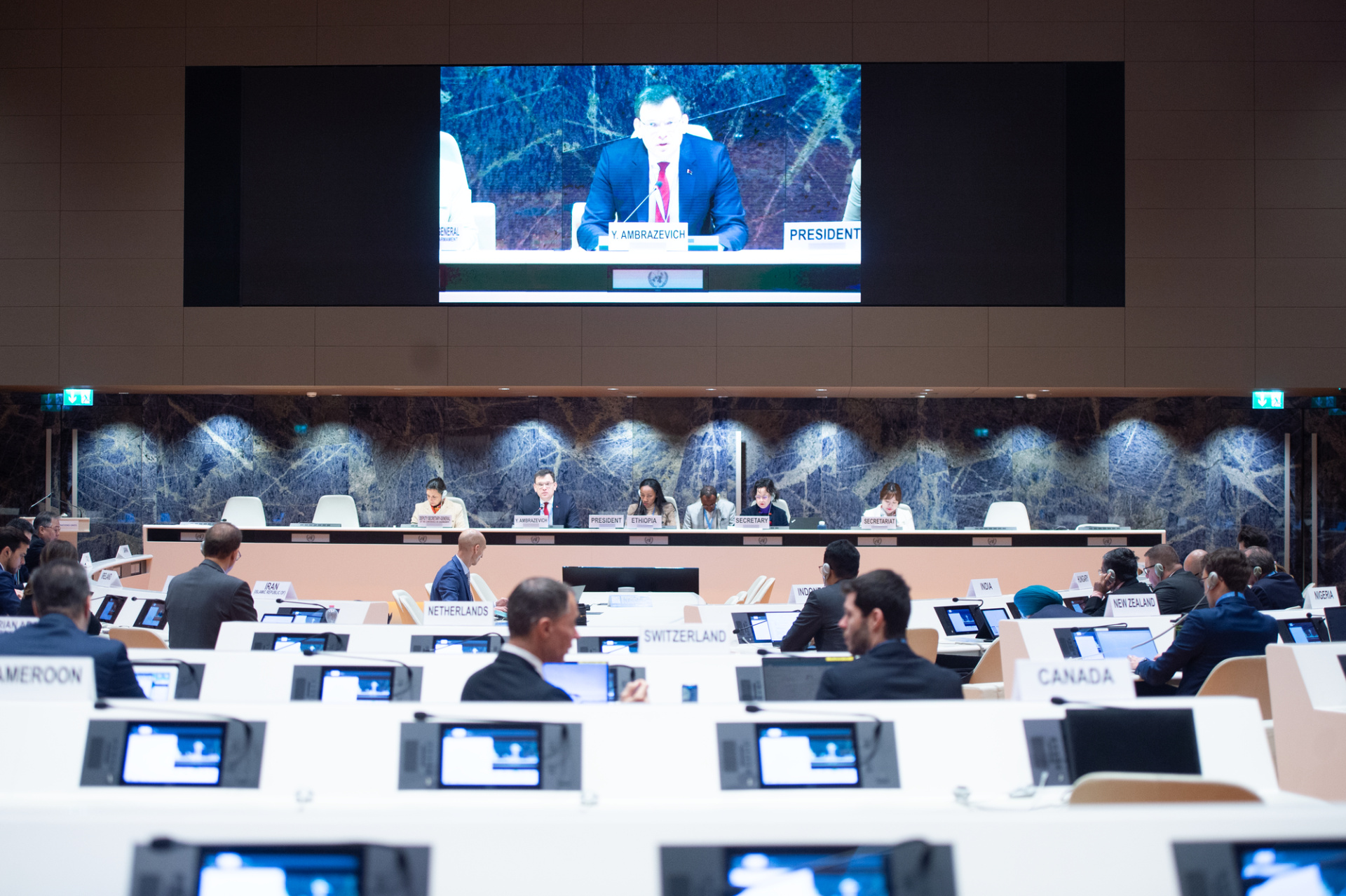 A wide shot of the conference room where participants of the Conference on Disarmament are currently listening to a speaker on screen. 