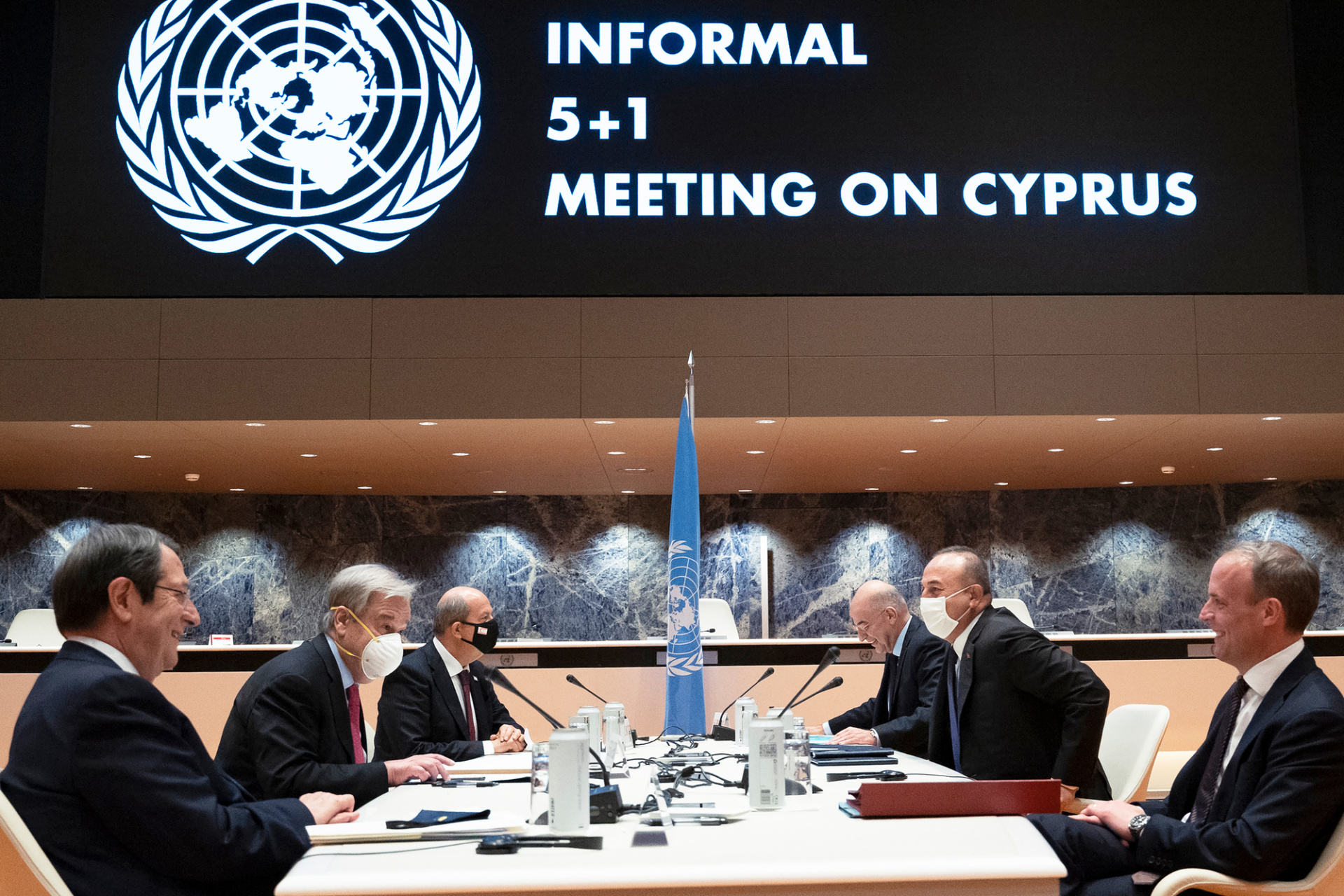 6 men sitting around a negotiating table with the UN flag standing in the middle between them. A screen in the background reads: "Informal 5+1 Meeting on Cyprus"