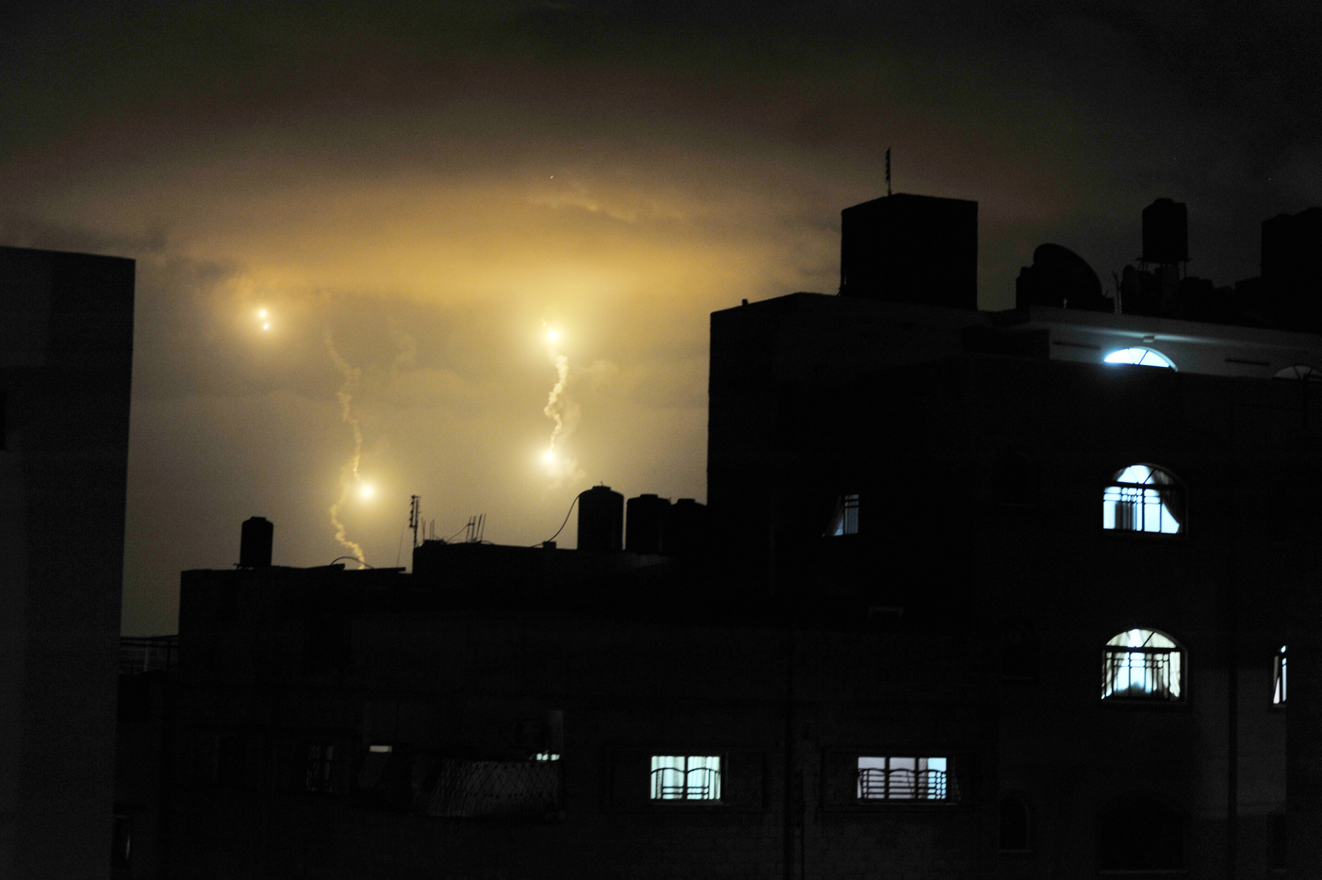 The shadows of a skyline at night are contrasted with bright lights in the sky coming from an air bombing on the city