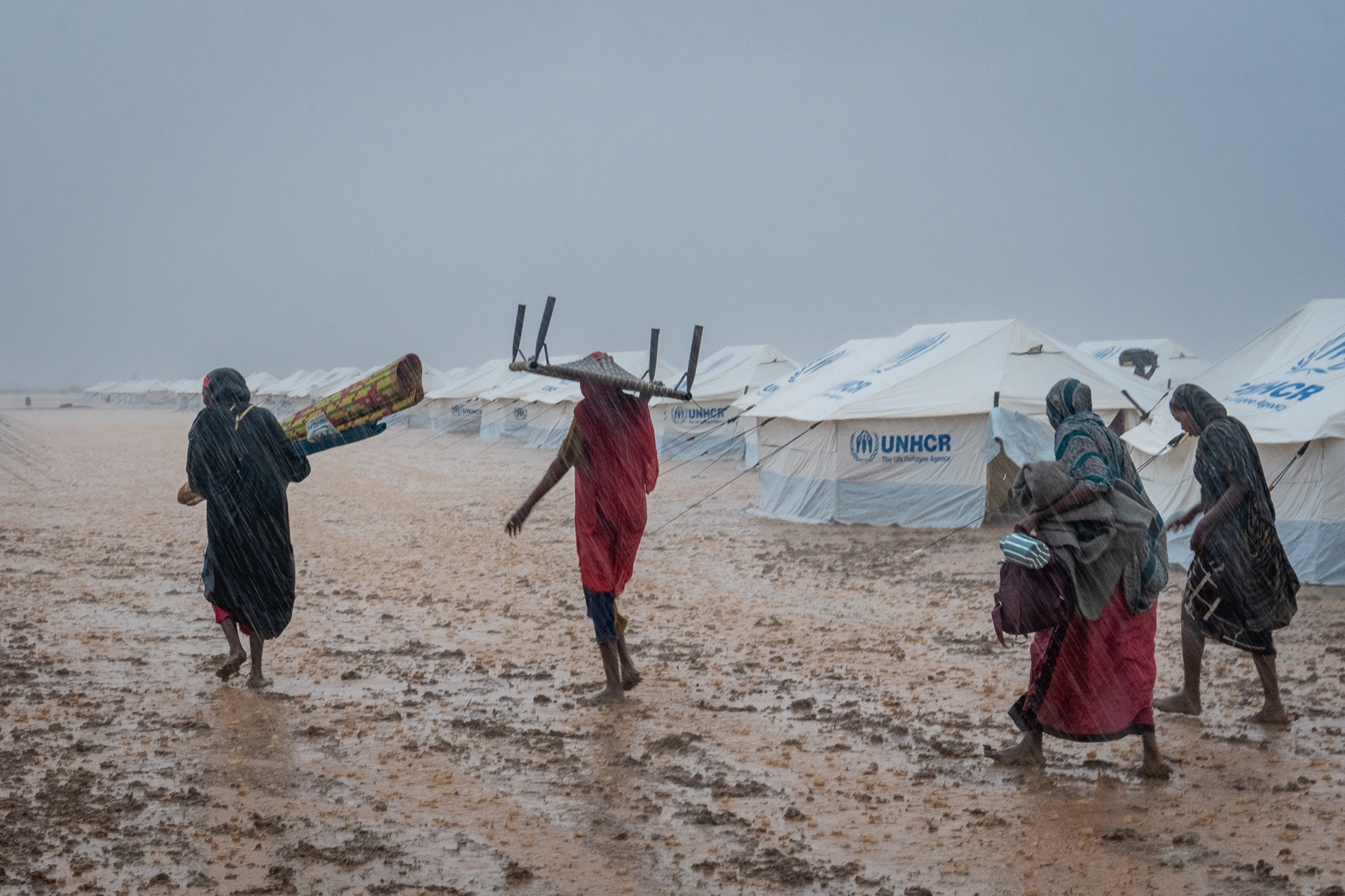 A group of refugee women make their way past tents through the rain, carrying furniture