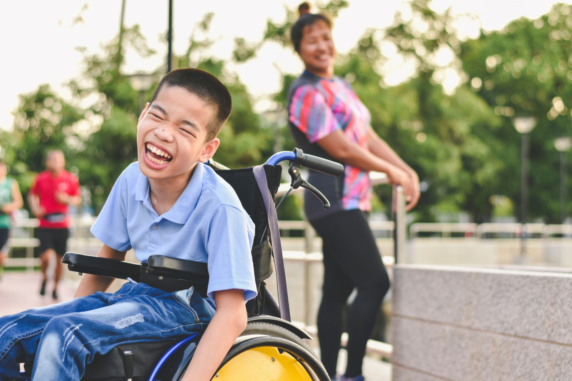 A laughing boy sits in a wheelchair in a school ground. A female adult is in the background, also smiling at the camera. 