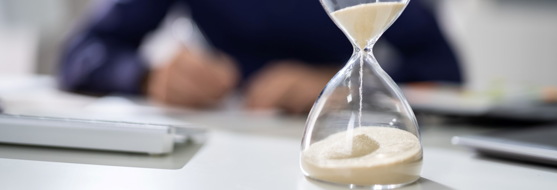 A sand glass in the foreground running empty while hands in the background, out of focus, write something on a paper.