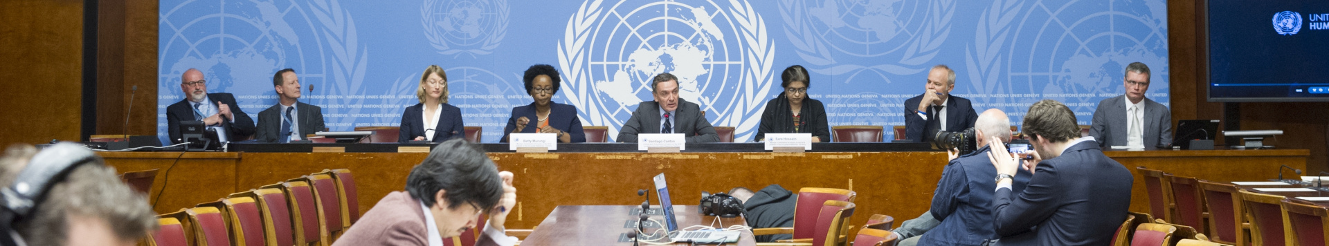 A press briefing at the Palais des Nations: Several UN staff sit in front of the UN logo, face to face with journalists.