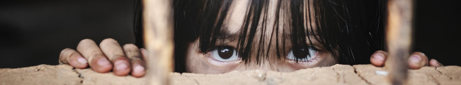 A girl peeps out from behind bars, only her eyes and hands are seen.